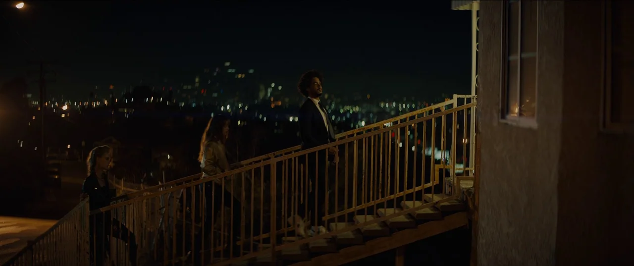 Night Teeth (2021) movie still: wide — A dark outdoor staircase overlooks a blurred city landscape illuminated by scattered night lights. Two people…