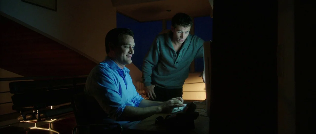 The Canyons (2013) movie still: medium — A dark, modern study illuminated by the harsh blue glow of a computer screen. Two men are huddled together…