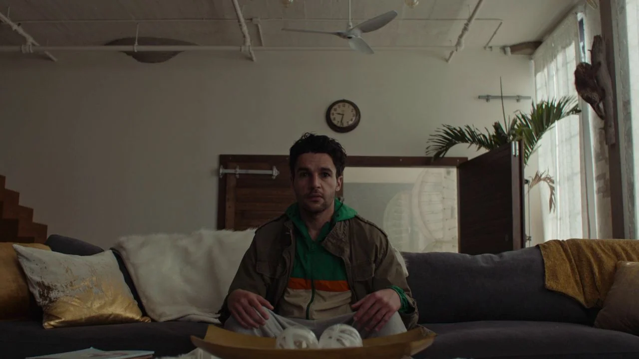 Possessor (2020) movie still: medium — A spacious living room features white walls, a ceiling fan, and minimalist wall decor. A man sits on a grey…
