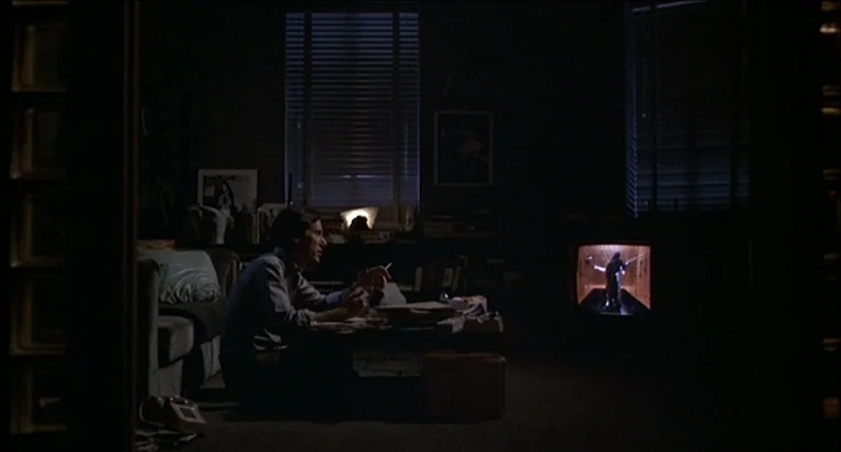 Videodrome (1983) movie still: medium — A dark, cluttered living room contains a man seated at a coffee table. The primary light source is a glowing…