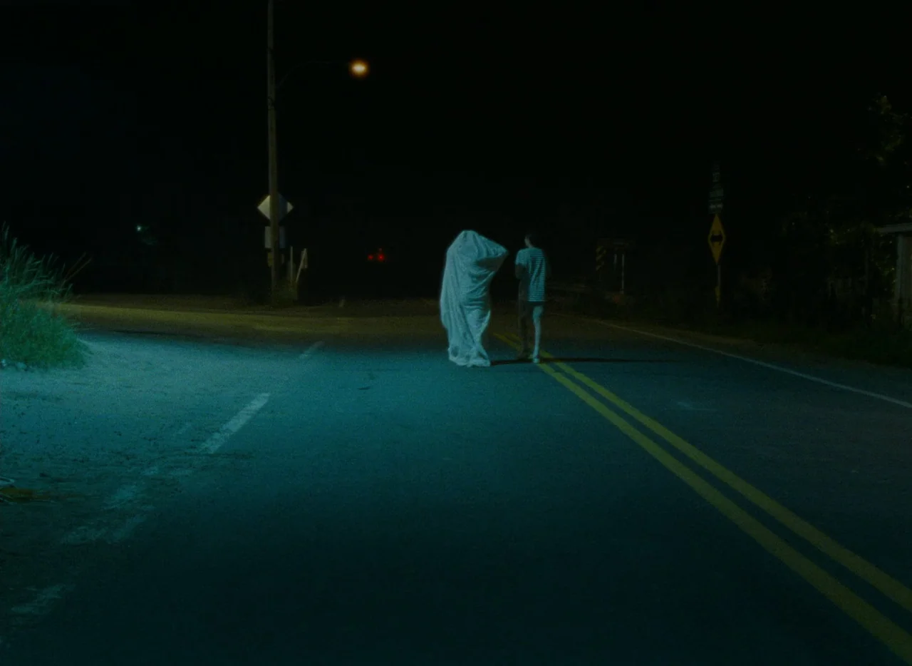 Falcon Lake (2022) movie still: wide — A quiet asphalt road at night marked by yellow lane dividers under streetlights. A person in a sheet costume…