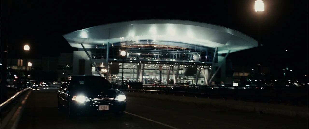 Zookeeper (2011) movie still: wide — A dark, modern airport terminal glows brightly against a pitch black night sky. A dark sedan moves along the…