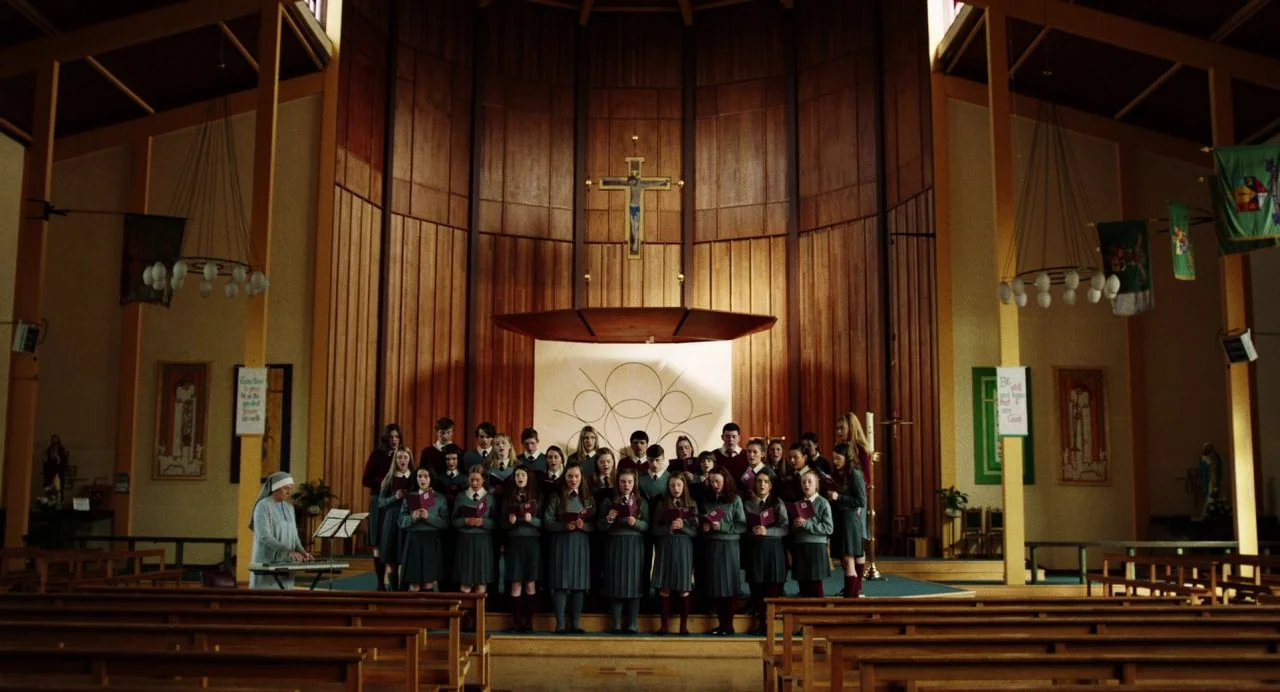 Dating Amber movie still: wide — A wooden church interior features a choir of students standing before a wall cross. The group stands on a small…