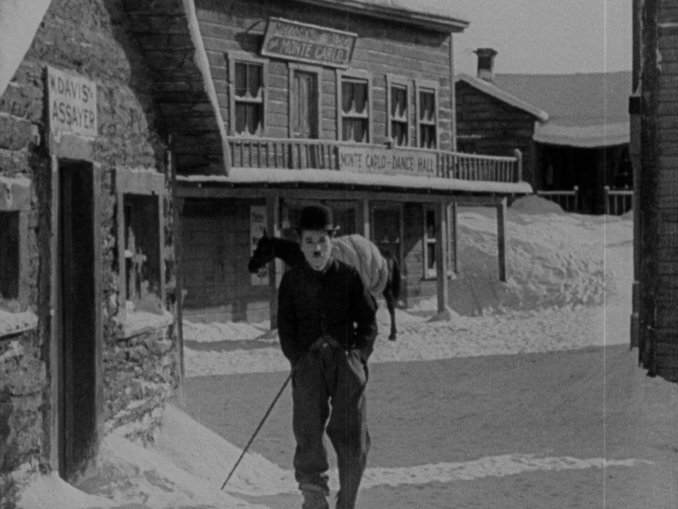The Gold Rush (1925) movie still: fullbody — A snowy Western street lined with rustic wooden buildings under clear daylight. A man with a cane walks…