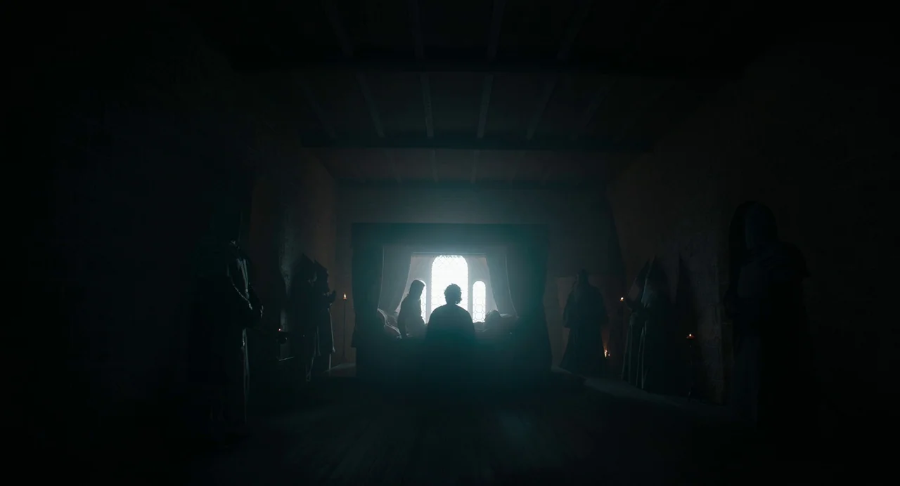 The Green Knight (2021) movie still: wide — A dark, vaulted stone room features a bed positioned directly in front of a window. Several cloaked…