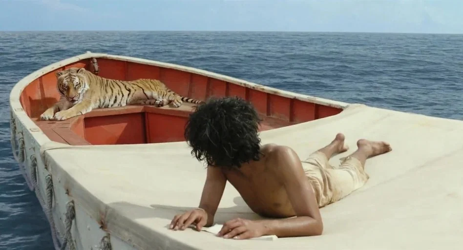 Life of Pi (2012) movie still: medium — A small orange lifeboat floats on the vast open ocean under a bright sky. A young man lies on the boat's…