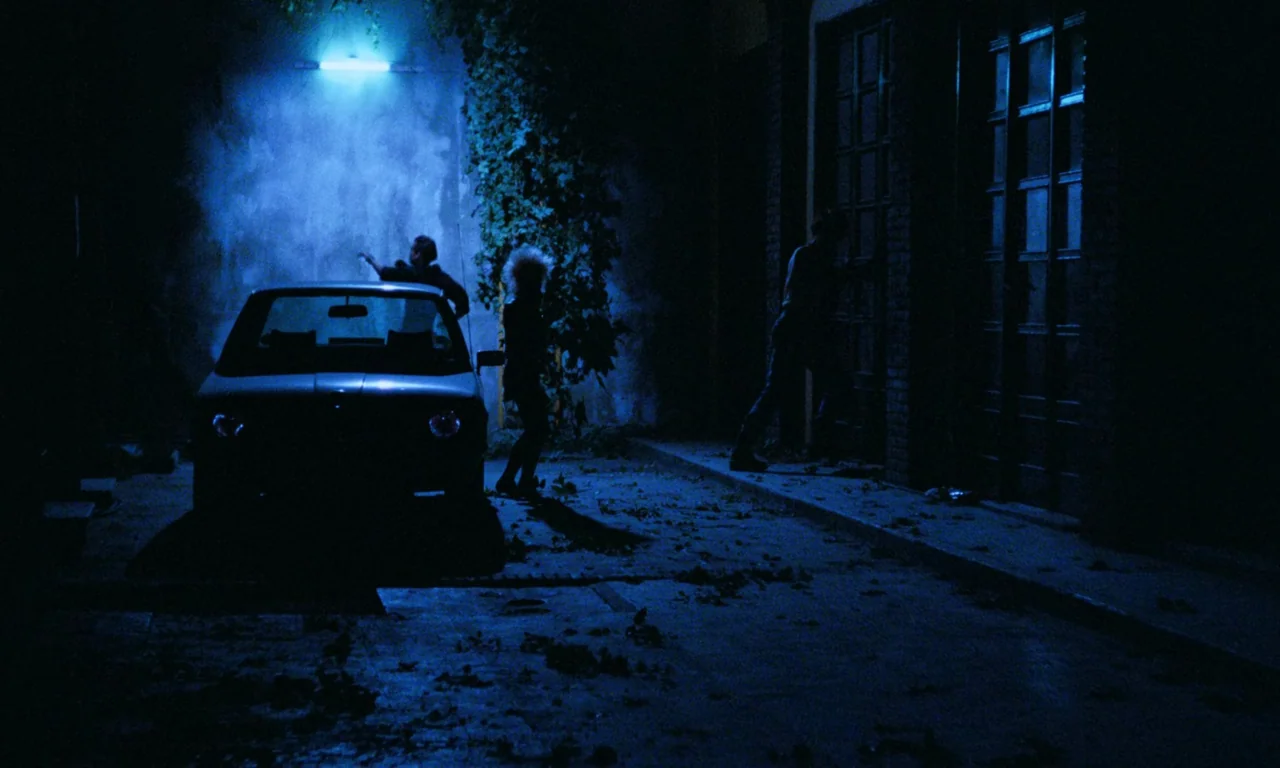 Demons (1985) movie still: wide — An urban alleyway at night illuminated by a single blue neon sign. Three figures move away from a brick building…