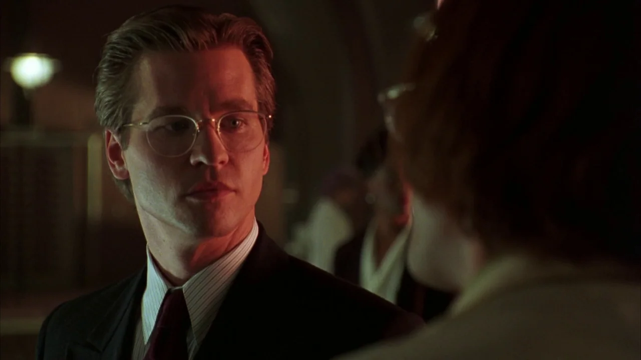 Batman Forever (1995) movie still: closeup — An interior space features a man listening to someone during an evening encounter. The man faces…