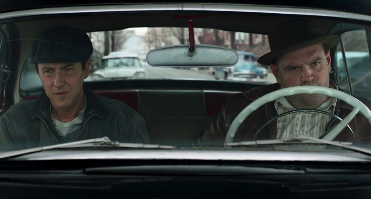 Motherless Brooklyn (2019) movie still: medium — Two men sit inside a 1950s sedan on a suburban city street. They occupy the front seats of the…