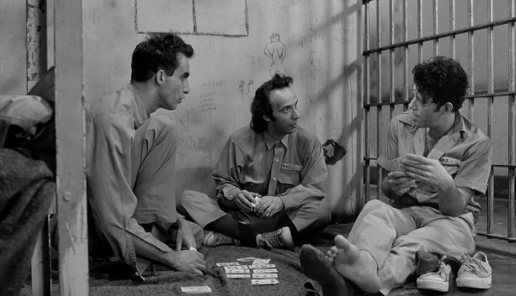 Down By Law (1986) movie still: fullbody — A small room features concrete walls covered in crude sketches and graffiti. Three men in uniform clothes…