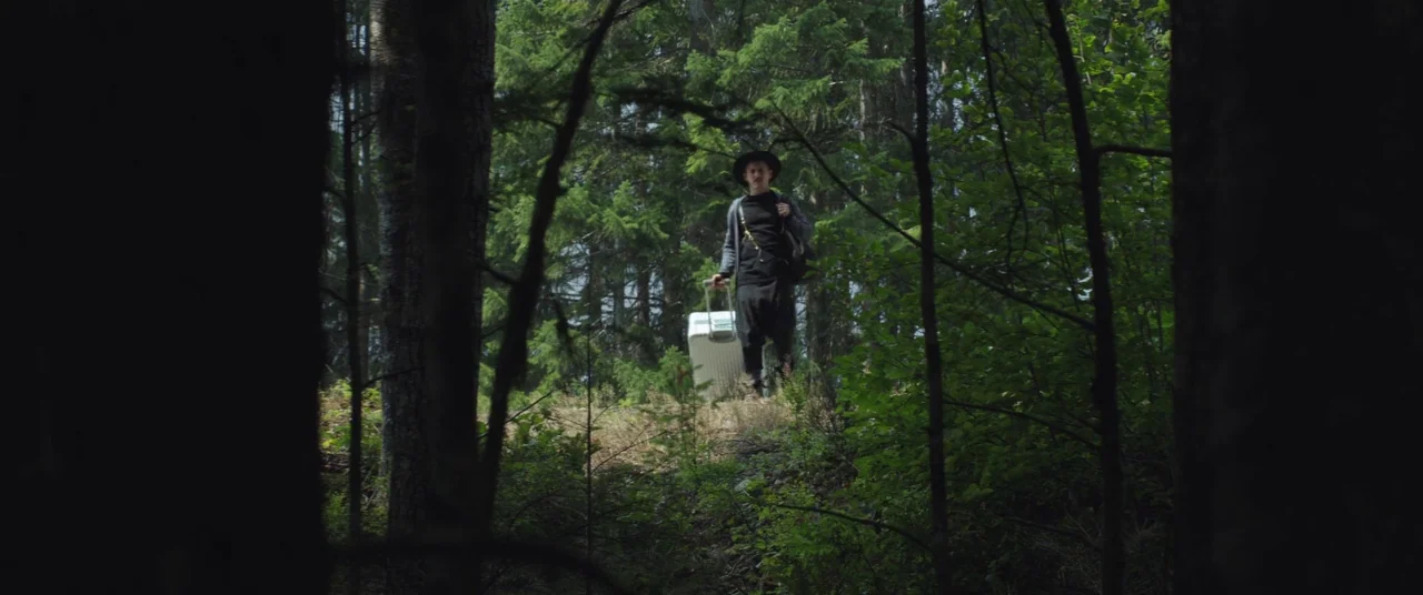 Come To Daddy (2019) movie still: wide — A dense patch of trees creates a natural frame around a person walking. A man dressed in black pulls a…