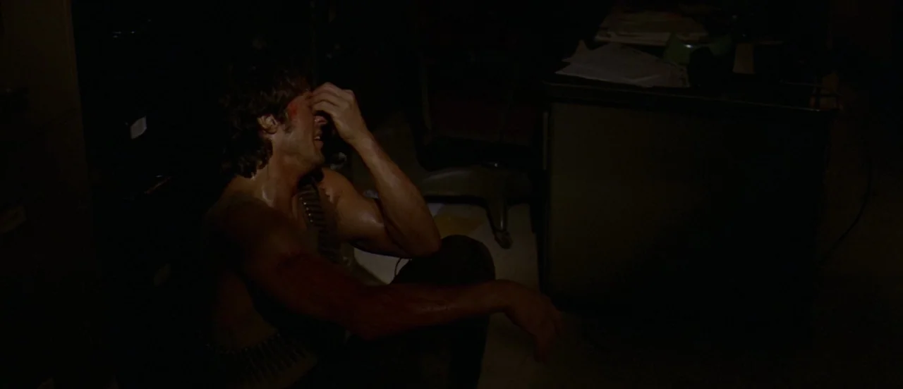 First Blood (1982) movie still: medium — A dark, dimly lit room contains a metal desk with scattered paperwork. A muscular man in a dark shirt sits…