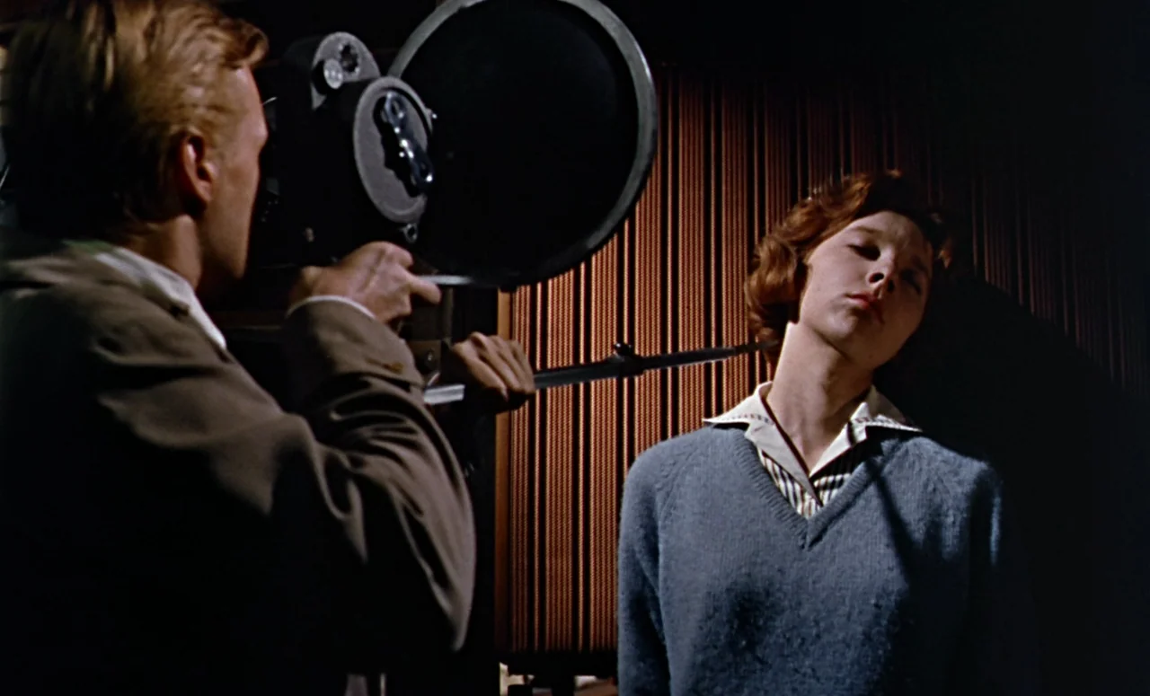 Peeping Tom (1960) movie still: medium — A professional recording space features vertical fabric wall treatments and low light levels. A man films a…