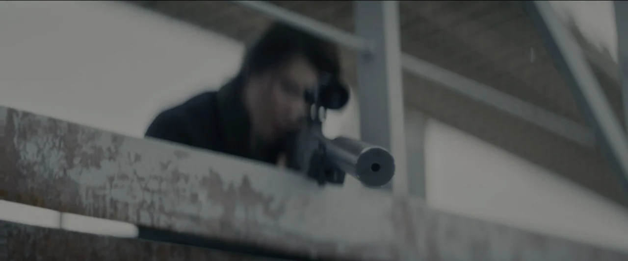 Kate (2021) movie still: medium — An industrial catwalk made of weathered metal overlooks an open structural space. A person in a dark jacket aims a…