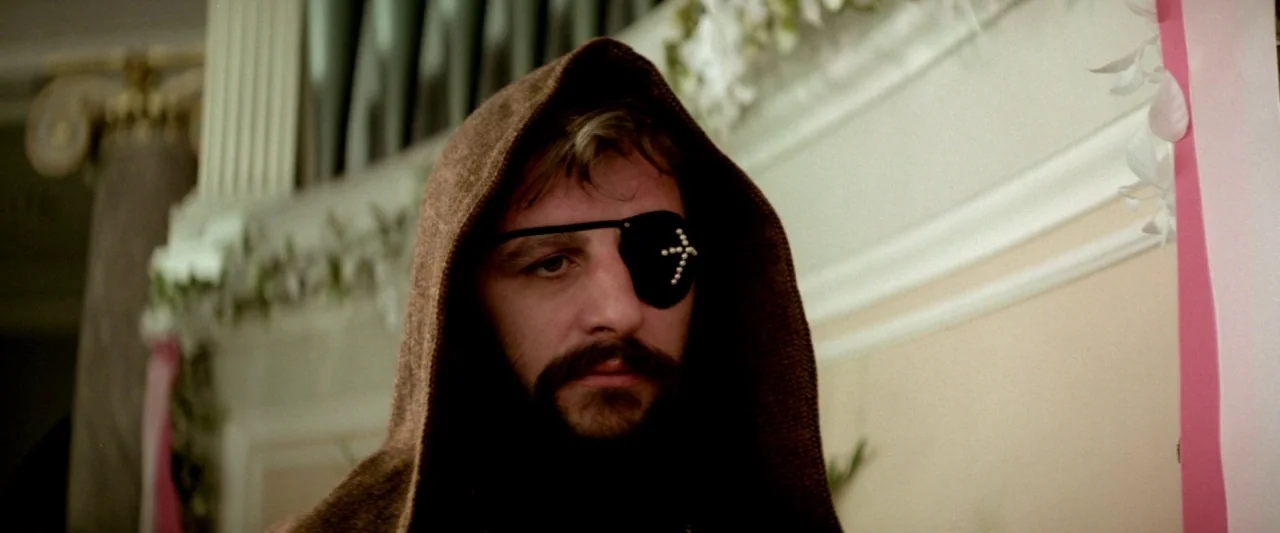 Lisztomania (1975) movie still: closeup — A man wearing a hooded cloak stands before a wall with draped fabric. The subject stares directly toward…