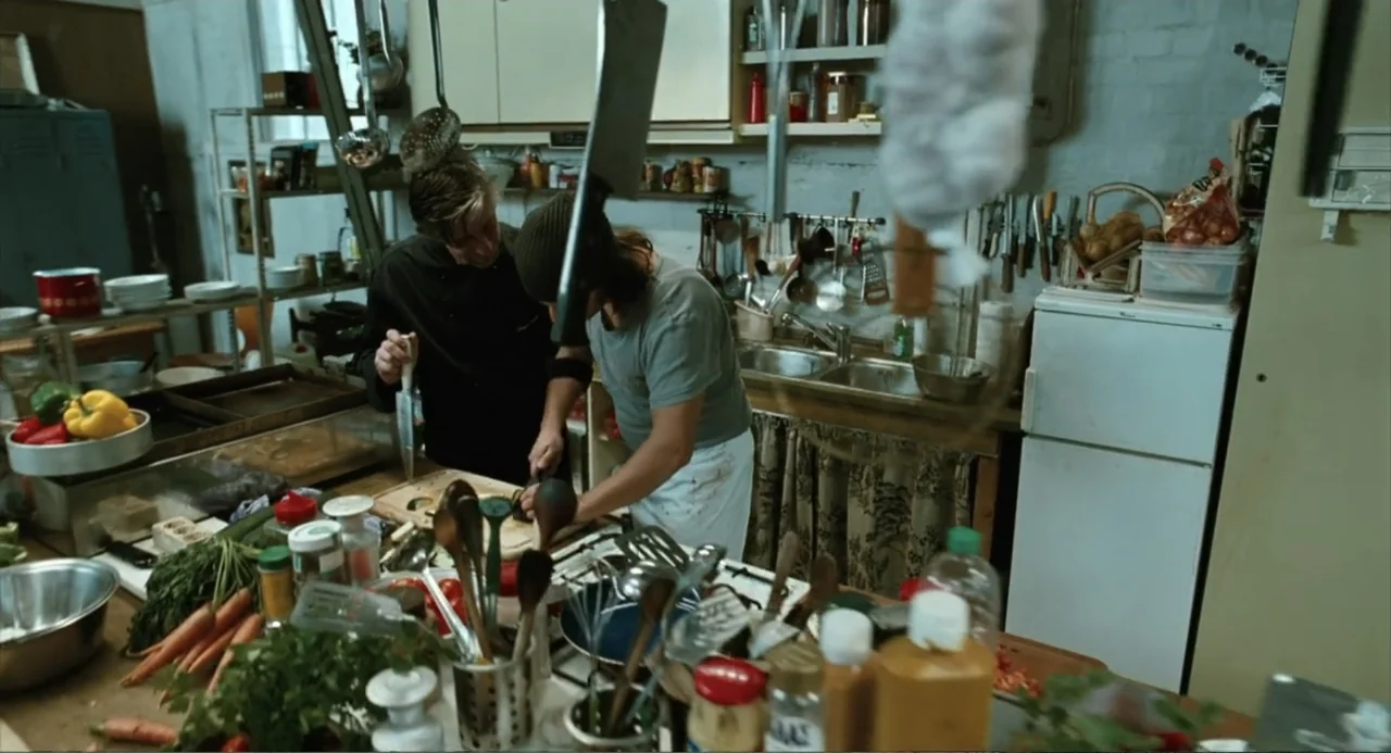 Soul Kitchen (2009) movie still: medium — A cluttered professional workspace filled with cooking equipment and fresh produce on counters. Two men in…