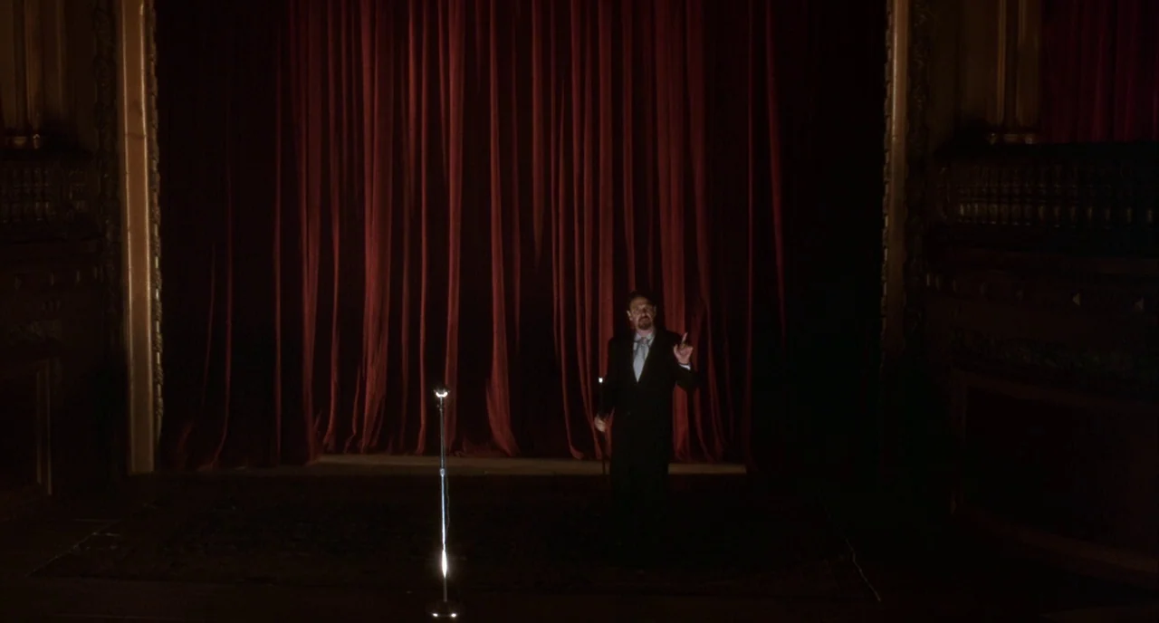 Mulholland Drive (2001) movie still: wide — A dimly lit theater stage features a large, dark red velvet curtain backdrop. A lone man in a suit…
