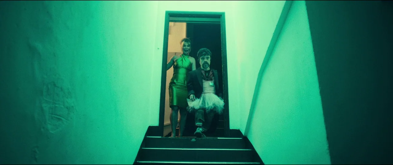 The Toxic Avenger (2023) movie still: fullbody — A dark stairwell is flooded with vibrant green light coming from an overhead fixture. A man in a…