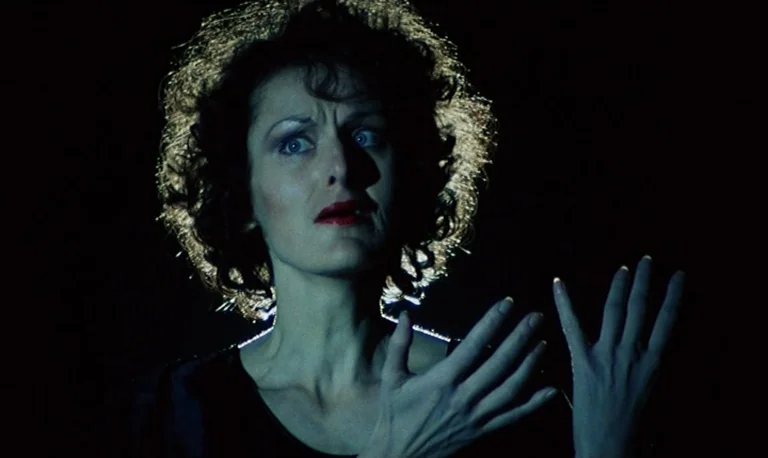 The Rose King (1986) movie still: closeup — A woman stands in a completely dark space illuminated only by a stark spotlight. She holds her hands up…