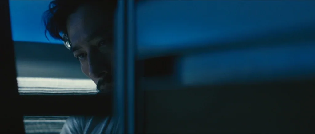 Sunshine movie still: closeup — A man looks at a screen in a dark room illuminated by blue light. The edge of a vertical partition divides the frame…