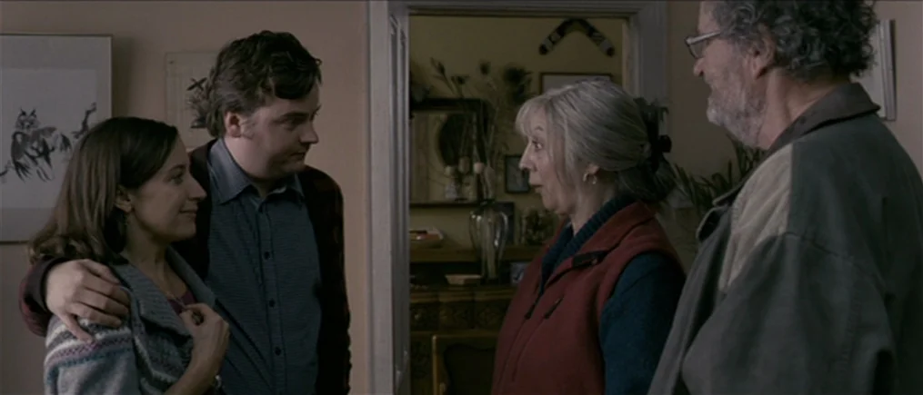 Another Year (2010) movie still: medium — A residential hallway with soft beige walls and simple artwork hanging on the side. A young couple stands…