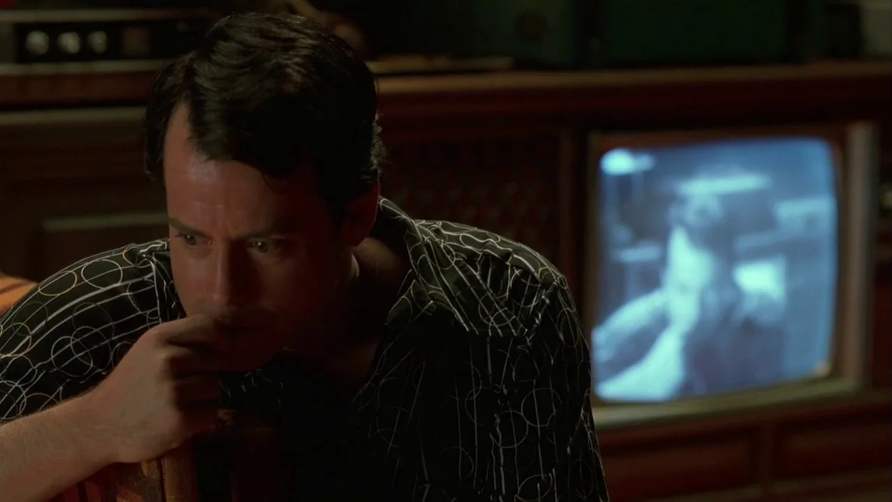 Auto Focus movie still: medium — A dimly lit room features a man sitting before a wooden entertainment console. A glowing television screen displays…