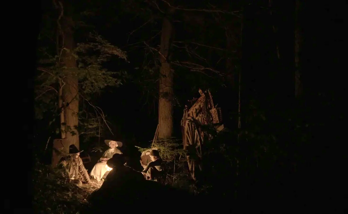 The Witch (2015) movie still: wide — A group of people sits around a fire in a dense, dark forest at night. They are positioned near a large wooden…