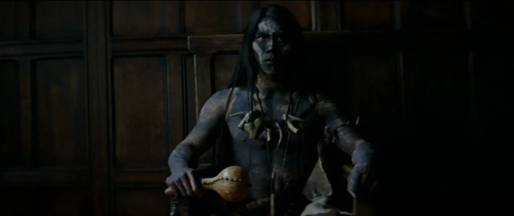 The New World (2005) movie still: medium — A man sits in a dimly lit room featuring wood-paneled walls. He holds a traditional gourd rattle while…