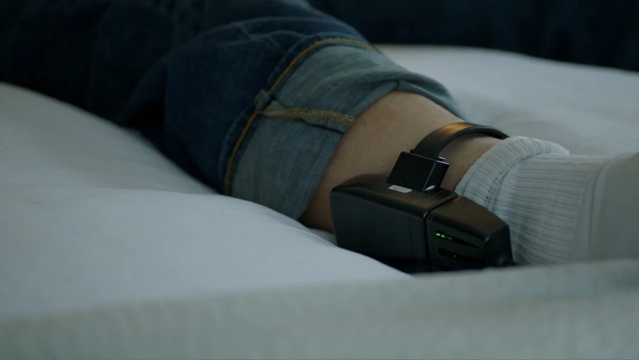 What Happened at Fells Acres? (2023) movie still: closeup — A person's lower leg rests upon a white fabric surface. An electronic tracking monitor…