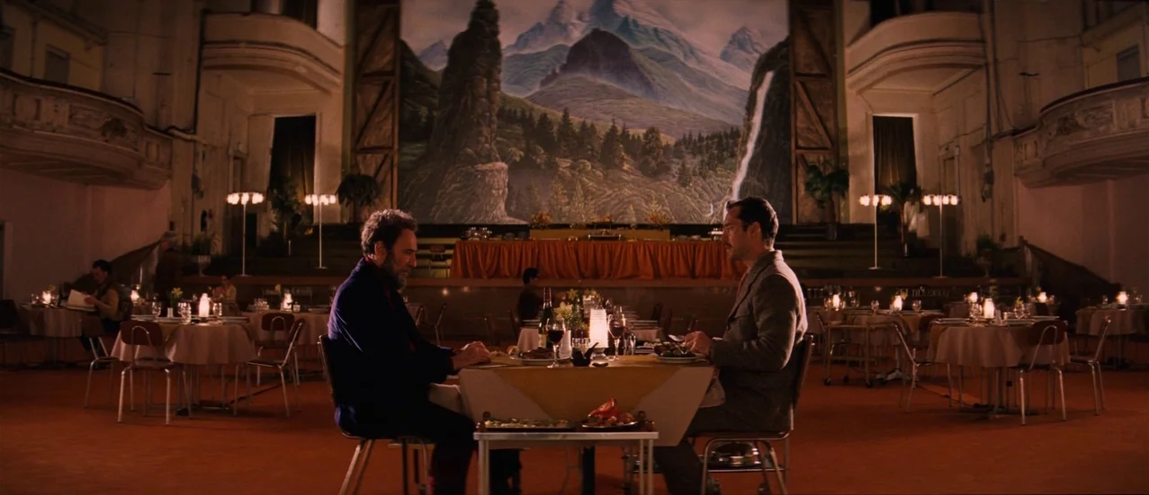 The Grand Budapest Hotel (2014) movie still: wide — A spacious dining hall features a large mountain mural backdrop and multiple circular tables.…