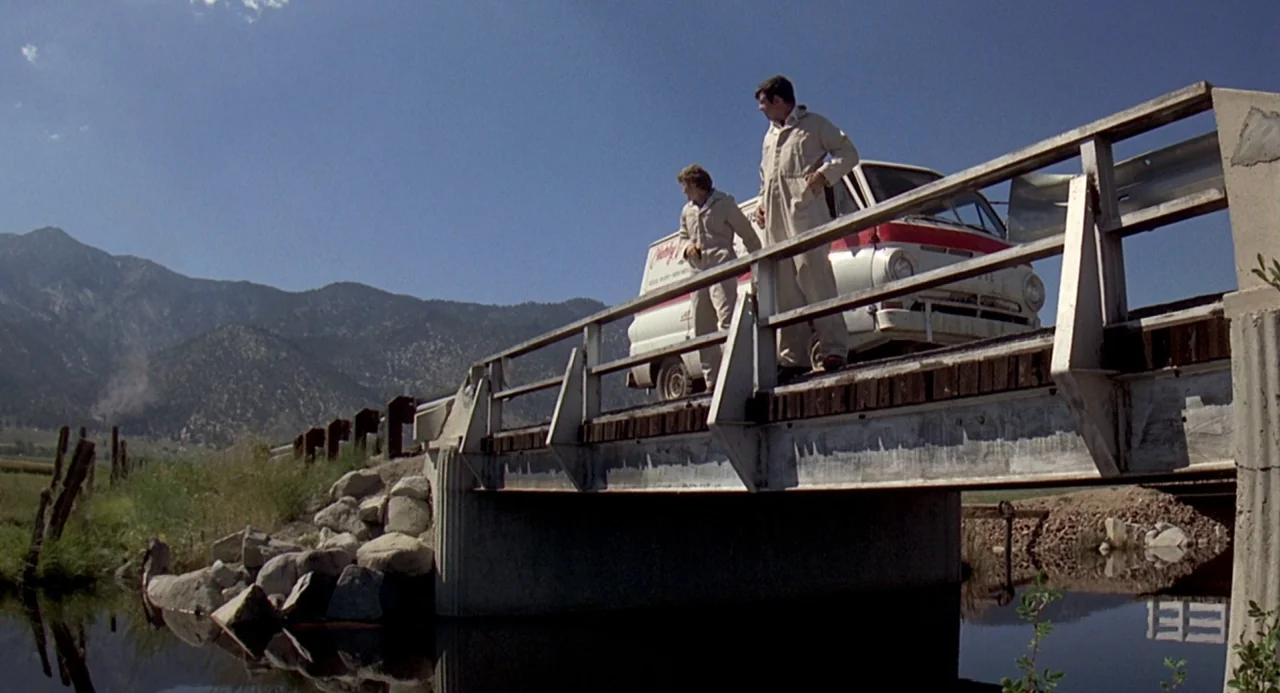 Charley Varrick (1973) movie still: wide — A wooden and steel bridge spans a calm stream in a mountainous rural landscape. Two men in work jumpsuits…