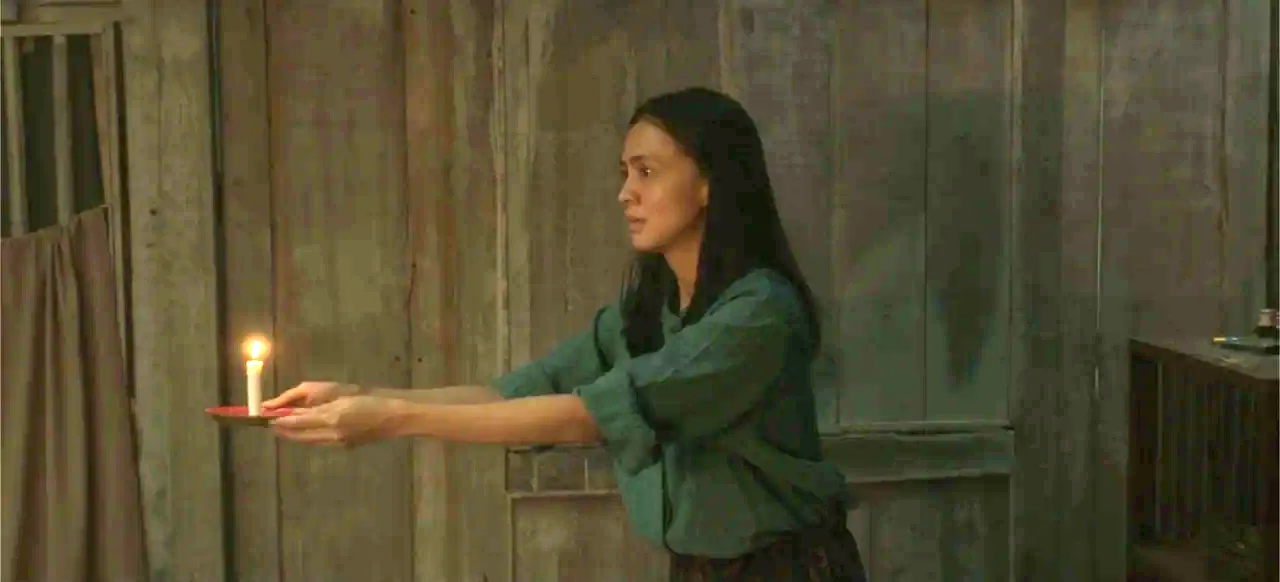 A Thousand Days (2023) movie still: medium — A dimly lit room with weathered vertical wooden plank walls. A young woman stands while holding a small…