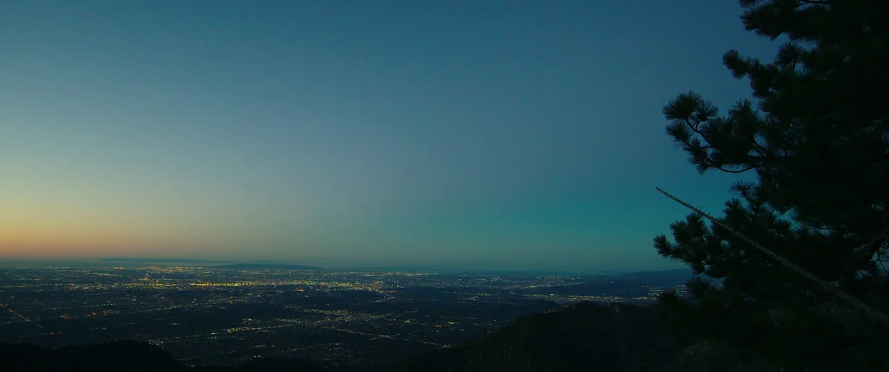 Nightcrawler (2014) movie still: establishing — A dark, elevated mountain ledge looks out over a sprawling city at twilight. Sparse pine branches…