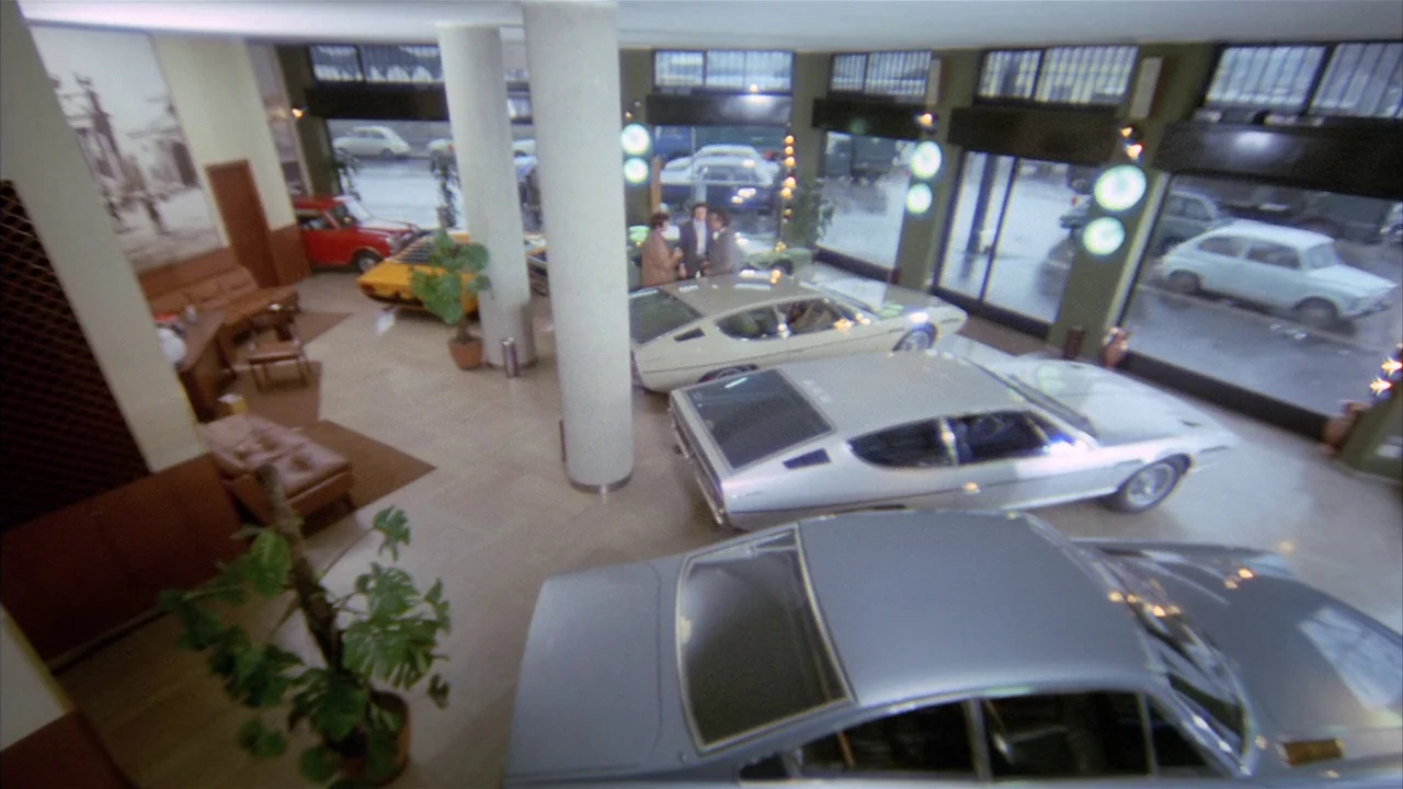 Death Occurred Last Night (1970) movie still: wide — A spacious, brightly lit showroom displays several vintage Italian sports cars on tiled floors.…