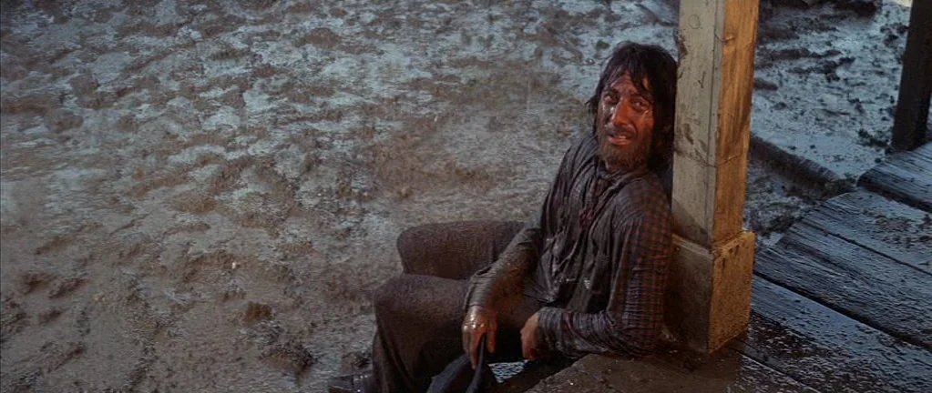 Little Big Man (1970) movie still: medium — An outdoor area covered in thick mud near a wooden building structure. A disheveled man sits on the wet…