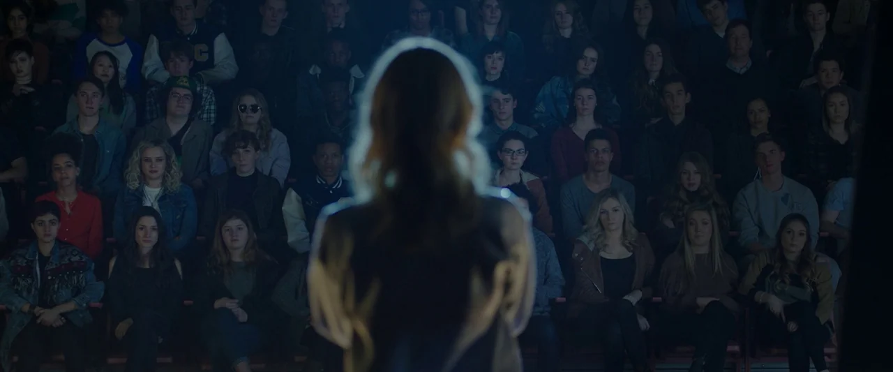 Spontaneous (2020) movie still: medium — A dark, densely packed auditorium is filled with rows of students watching a presentation. A woman stands…