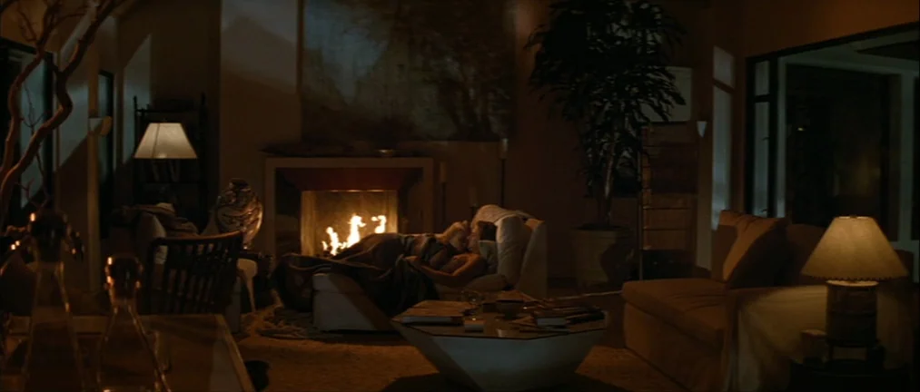 Basic Instinct (1992) movie still: wide — A spacious room features a fireplace flanked by a large potted plant and furniture. Two people rest on a…