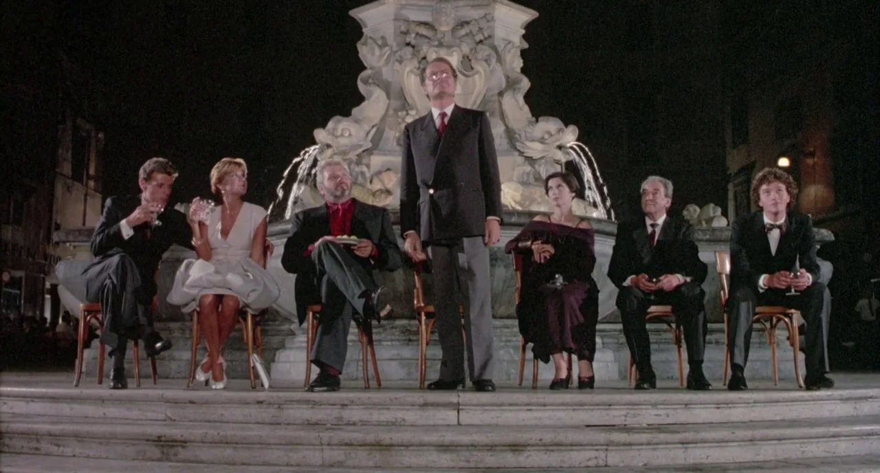 The Belly of an Architect (1987) movie still: wide — A group of formally dressed adults sit or stand on stone steps at night. A large, ornate stone…