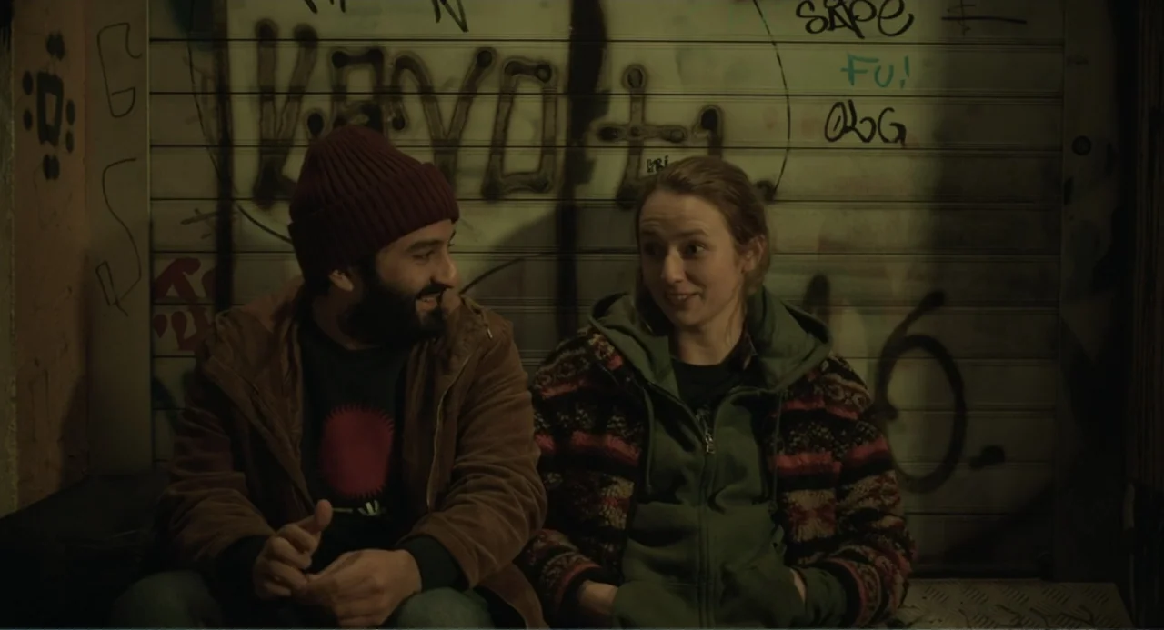 Ninjababy (2021) movie still: medium — A night scene shows a couple sitting against a metal shutter covered in graffiti. The pair faces each other…