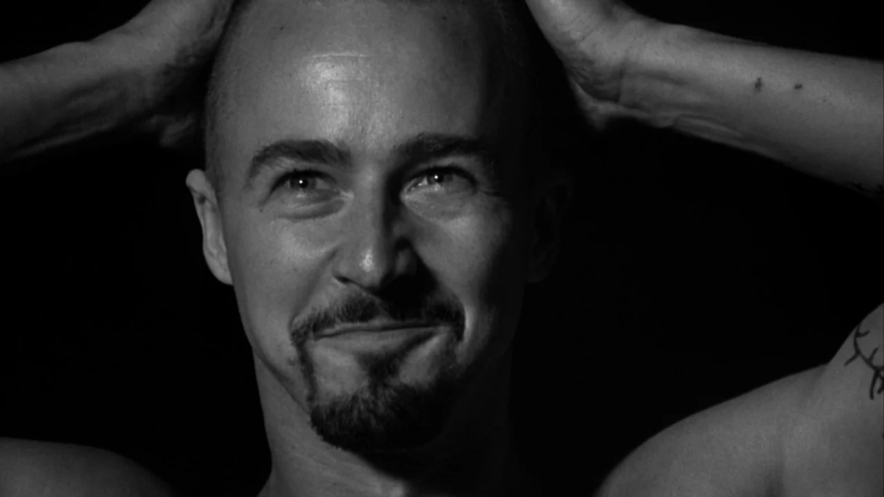 American History X movie still: closeup — A man stands against an pitch black background with his hands behind his head. The lighting highlights his…