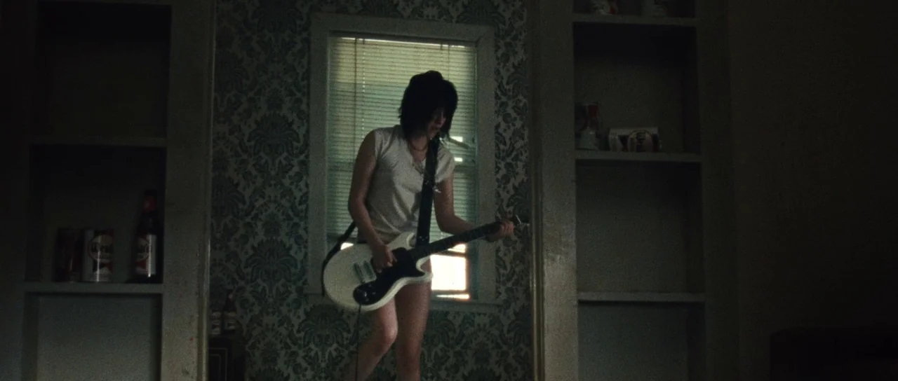The Runaways (2010) movie still: medium — A dark, dimly lit room features patterned wallpaper and built-in wooden shelving units. A young woman in a…