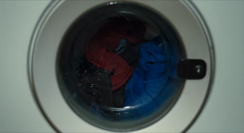35 Shots of Rum (2008) movie still: closeup — A close-up view shows the circular glass door of a front-loading washing machine. Wet, colorful…