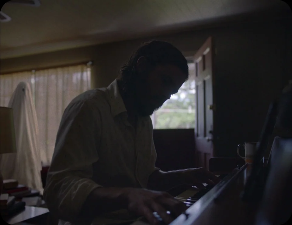 A Ghost Story (2017) movie still: medium — A dimly lit room contains a piano, a sheet-draped figure, and a window with curtains. A man with a beard…