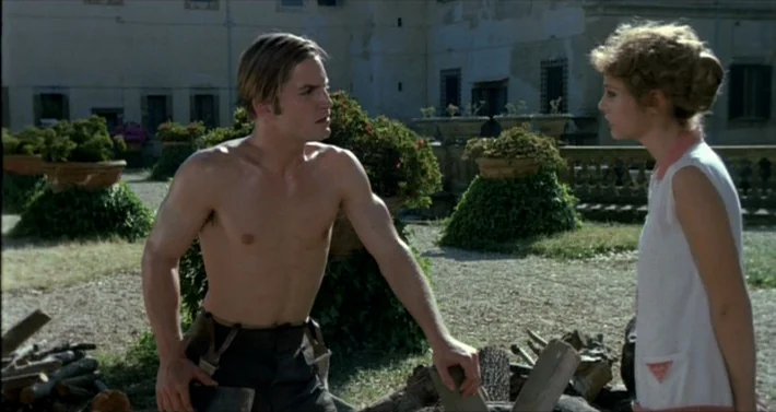 Blood For Dracula (1974) movie still: medium — A stone courtyard features ornamental bushes in large terracotta pots and a woodpile. A shirtless man…