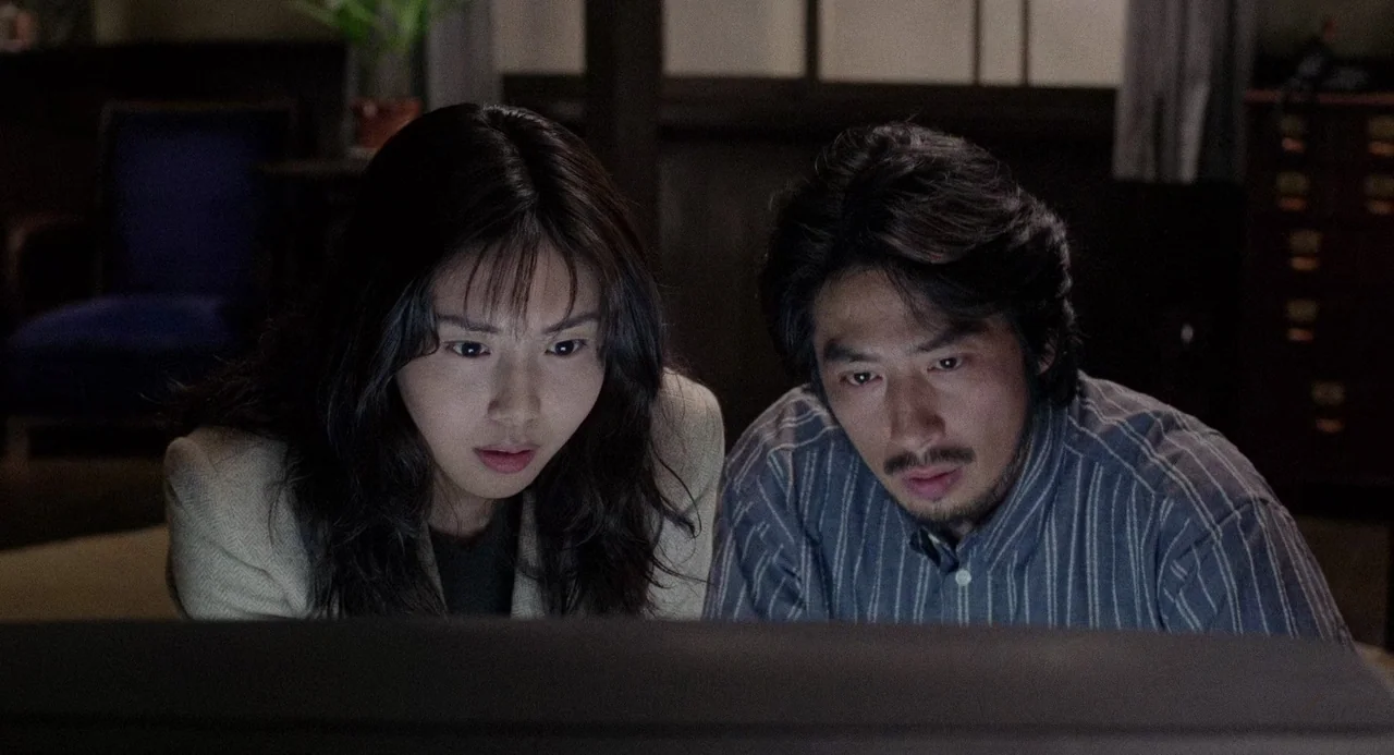 Ringu (1998) movie still: medium — A dark indoor space is dimly lit by the reflection of an unseen screen. An Asian man and woman stare intently at…