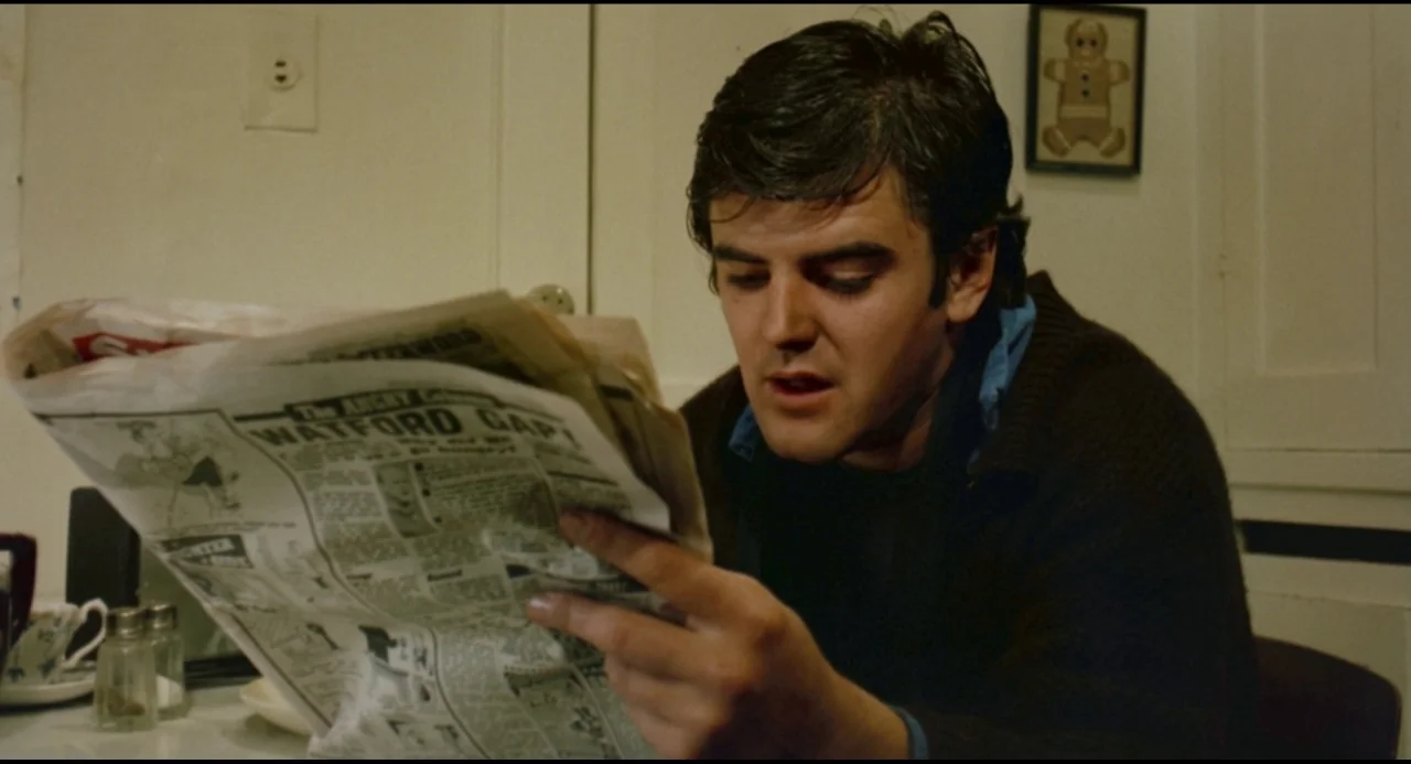 Olivia (1983) movie still: medium — A man sits at a table in a dimly lit room with plain walls. He holds a large newspaper with both hands while…