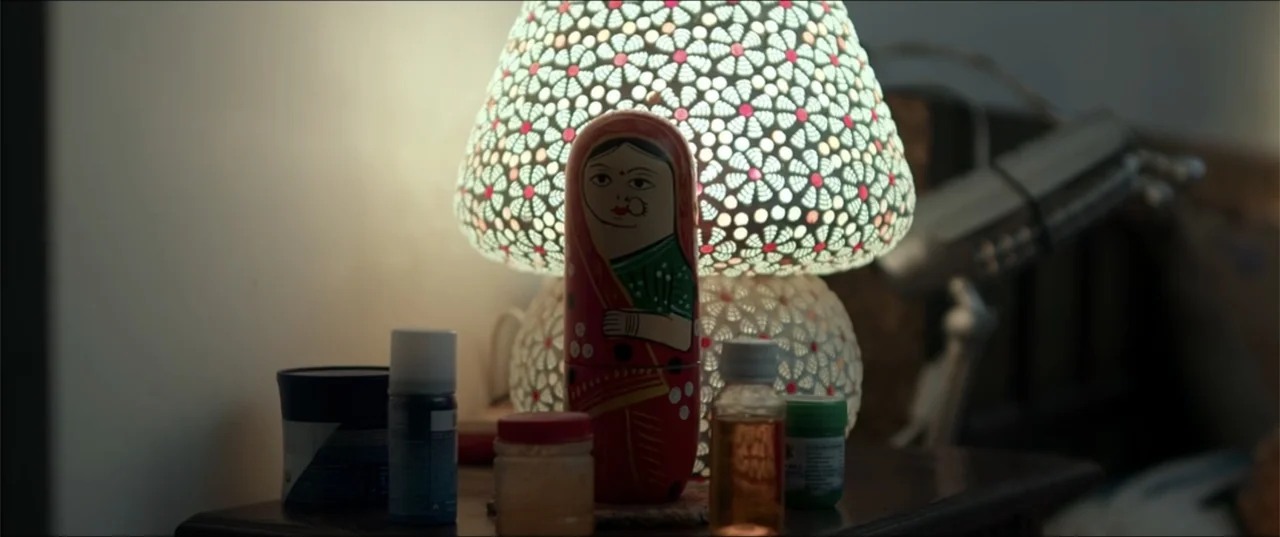 Cheekatilo (2026) movie still: closeup — A bedside table holds a decorative doll and several small cosmetic containers. The items are arranged in…
