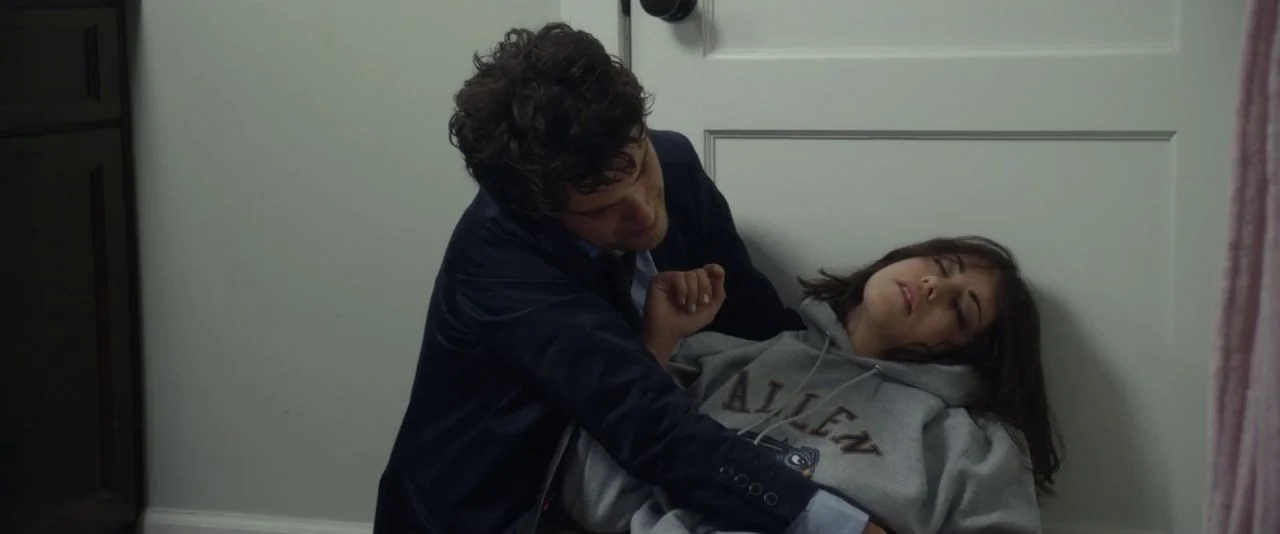 Night Owls (2015) movie still: medium — A quiet indoor hallway with a white door and wood flooring. A man leans over and holds a woman in a grey…