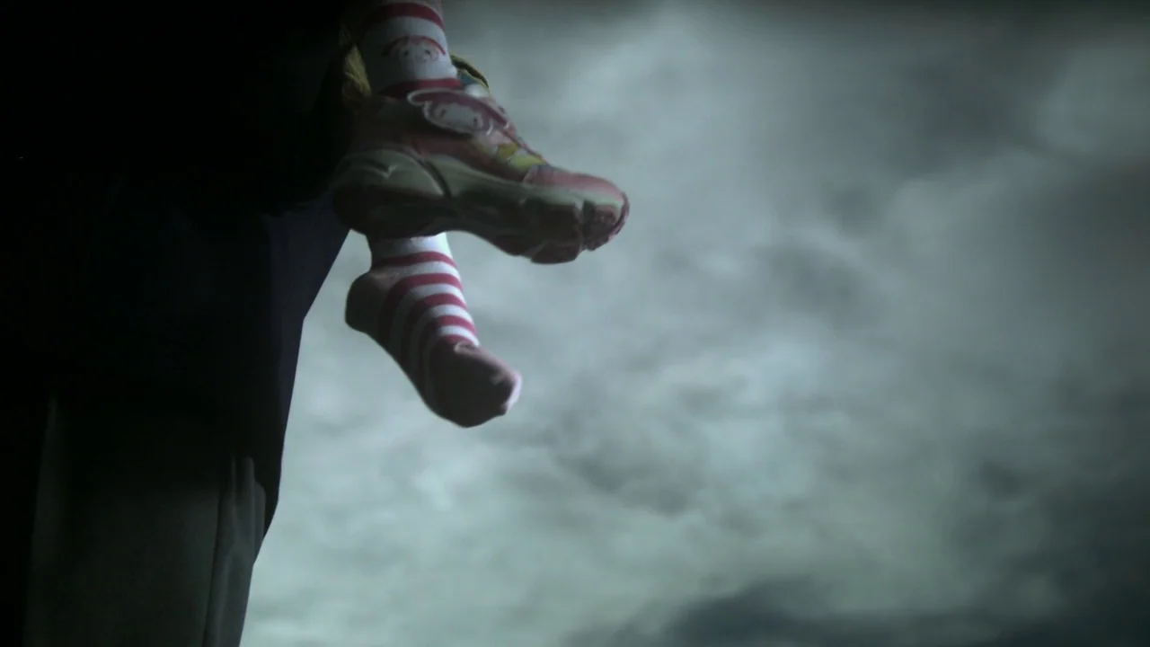 Confessions (2010) movie still: closeup — Child's legs in striped socks and sneakers dangle against a cloudy night sky. The background is a vast…