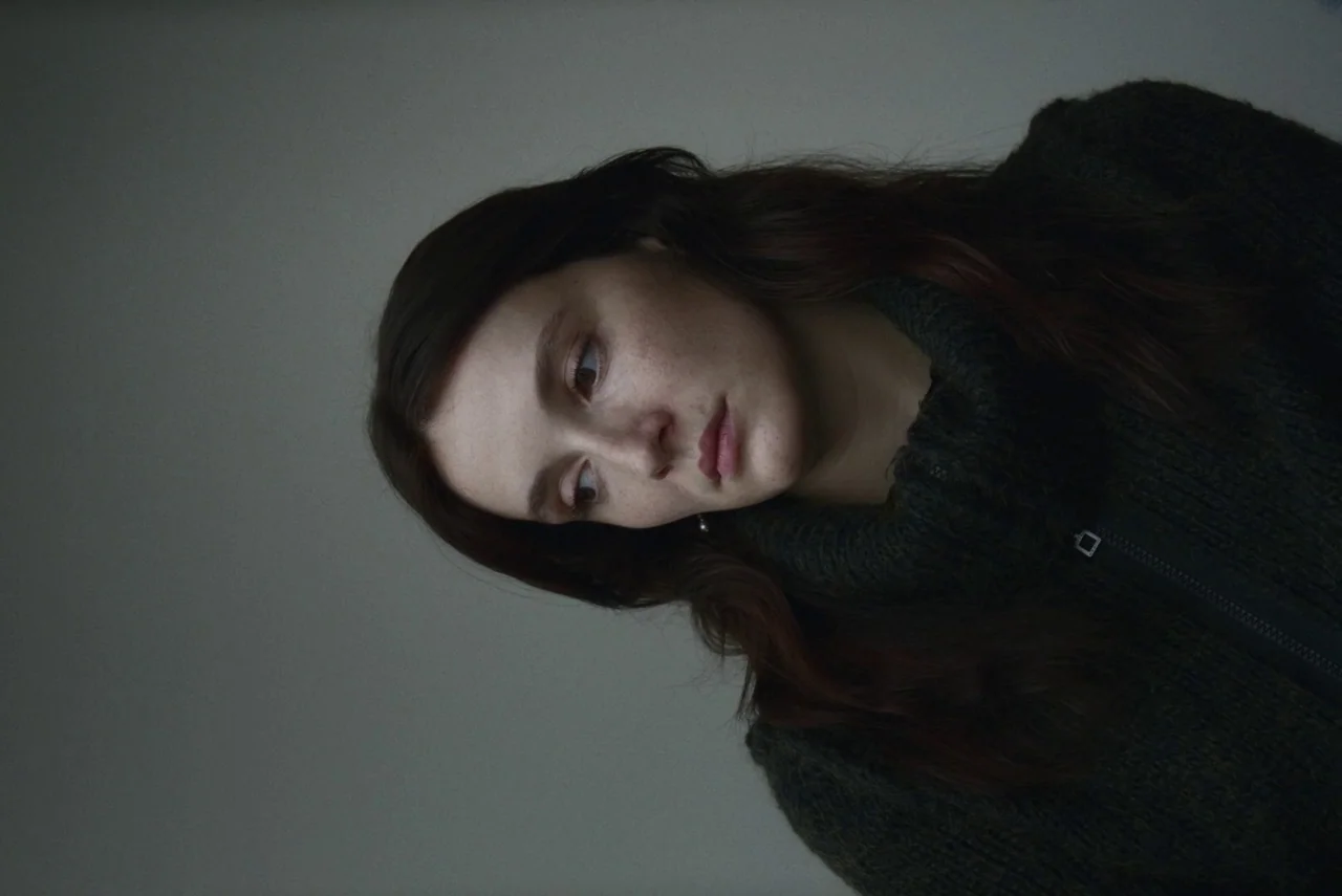 Red Rooms (2023) movie still: closeup — A plain gray wall serves as a backdrop for a portrait of a person. A woman with auburn hair wears a dark…